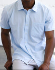 Man wearing a light blue shirt sitting against a white wall.