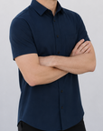 A man wearing a dark navy blue short-sleeve button-down shirt and black pants, standing with arms crossed against a plain light gray wall.